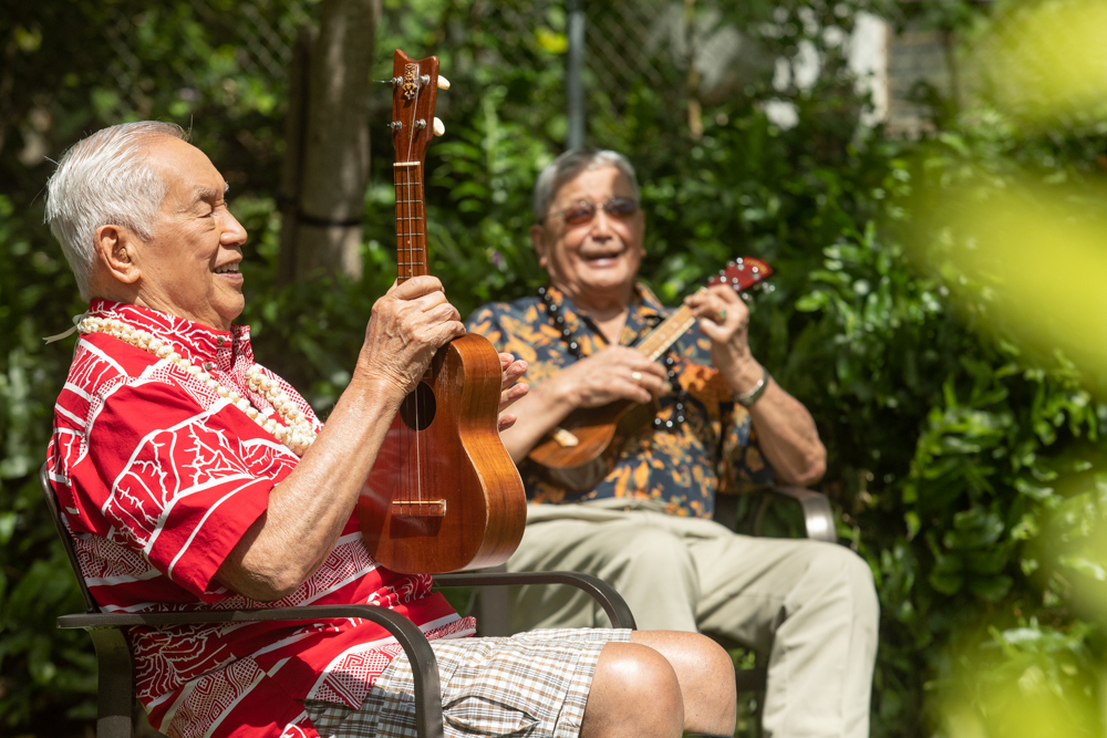 Two senior men laughing and playing ukuleles in a sunlit garden.