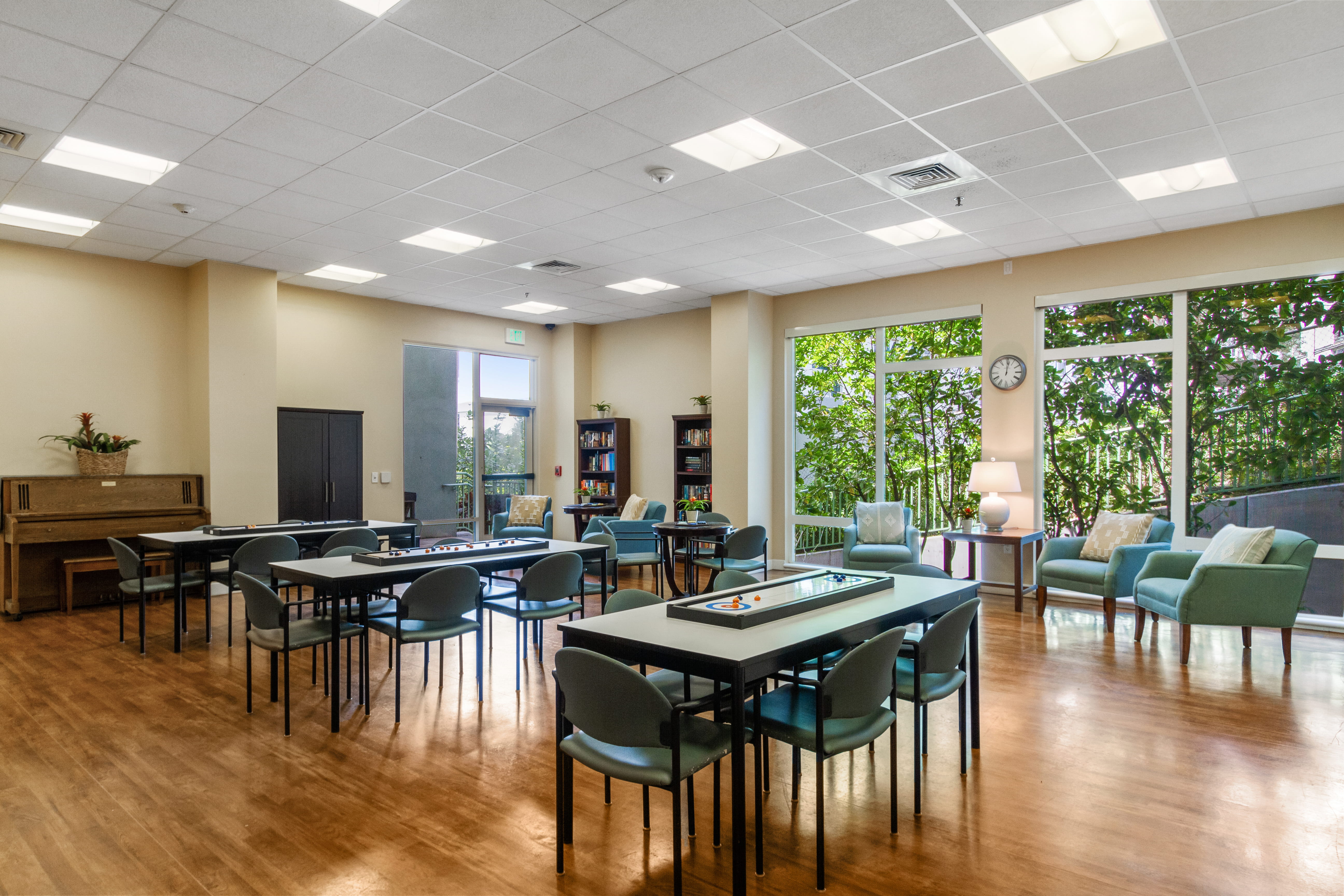 Large activity room with tables, a piano, and bright windows at The Plaza Assisted Living.