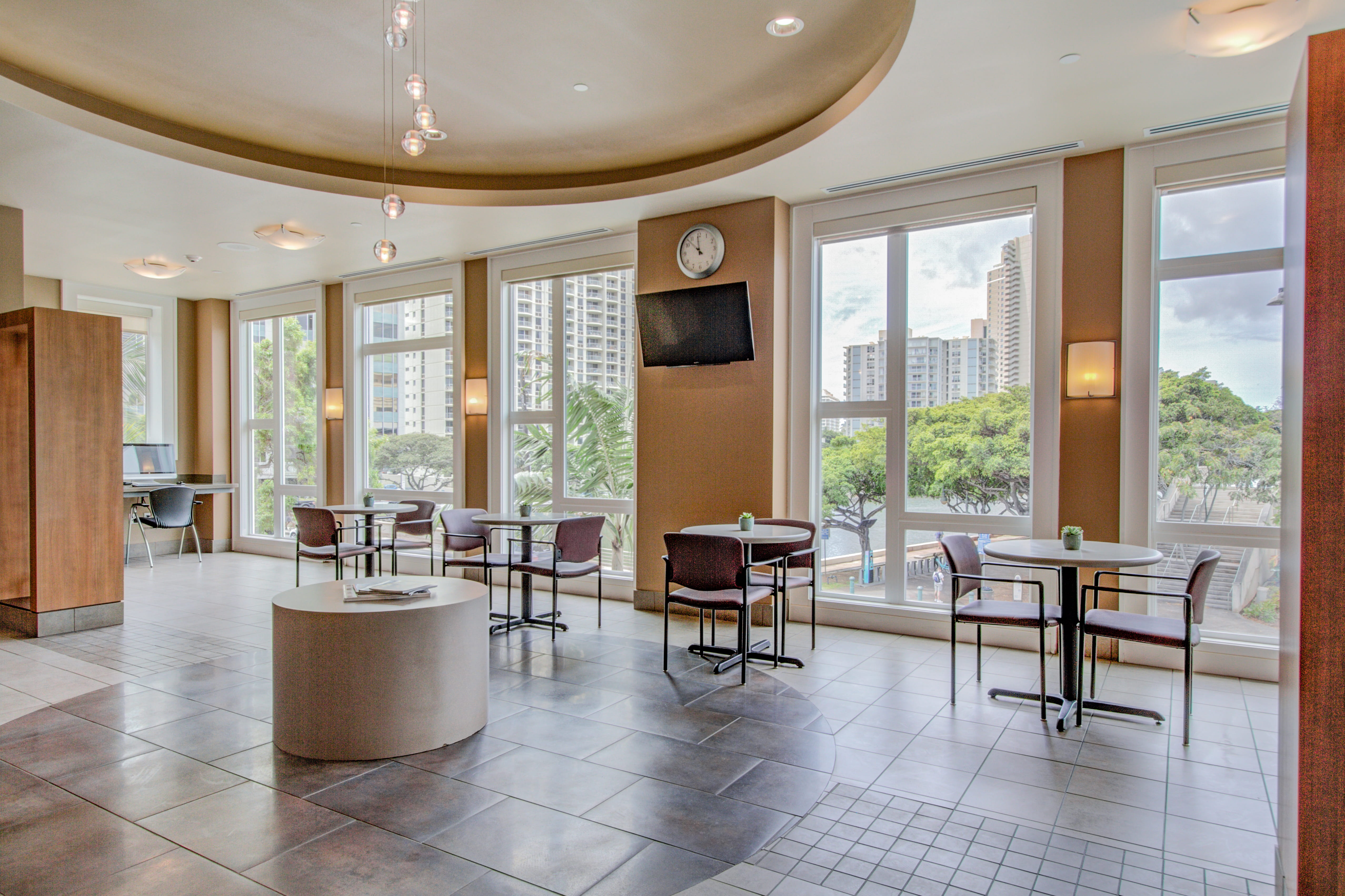 Sunlit bistro area with tables and windows overlooking Waikiki city trees.